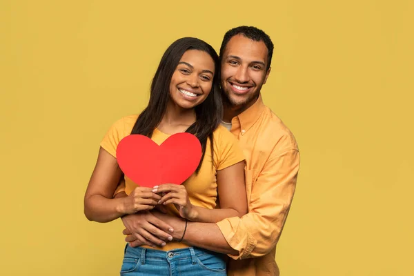 Romantic young black couple hugging, holding red paper heart on yellow studio background — Zdjęcie stockowe