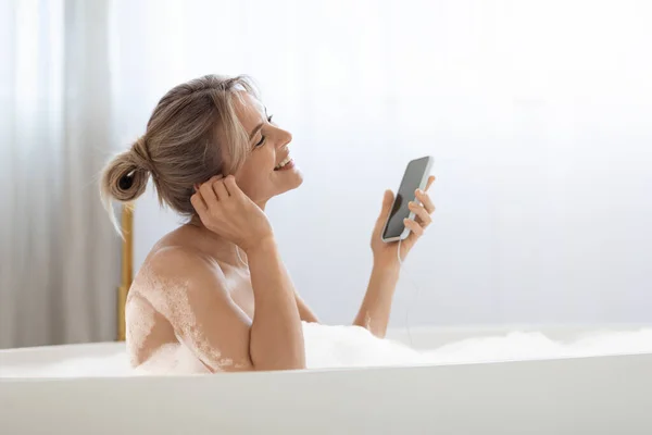 Mujer rubia positiva tomando un baño espumoso y escuchando música —  Fotos de Stock