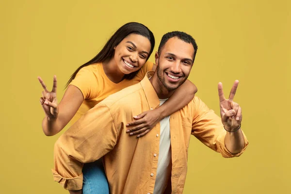 Handsome young African American man giving piggyback ride to his girlfriend, showing peace gesture on yellow background — Fotografia de Stock