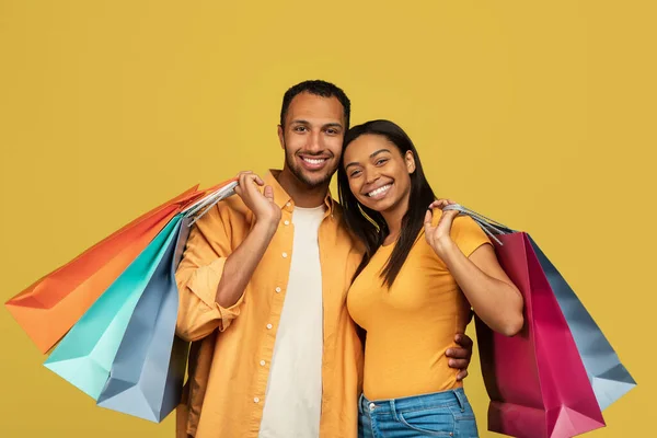 Happy young black couple with shopping bags smiling and looking at camera on yellow studio background — Photo