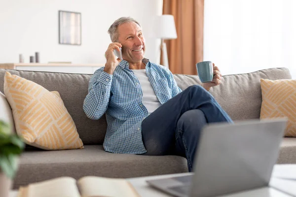 Χαρούμενος ώριμος άντρας που μιλάει στο κινητό και κάθεται στον καναπέ. — Φωτογραφία Αρχείου
