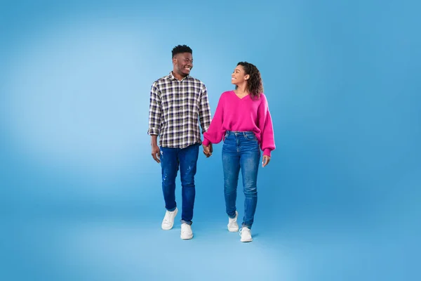 Happy millennial black couple walking, holding hands, looking at each other on blue studio background, full length — Stock fotografie