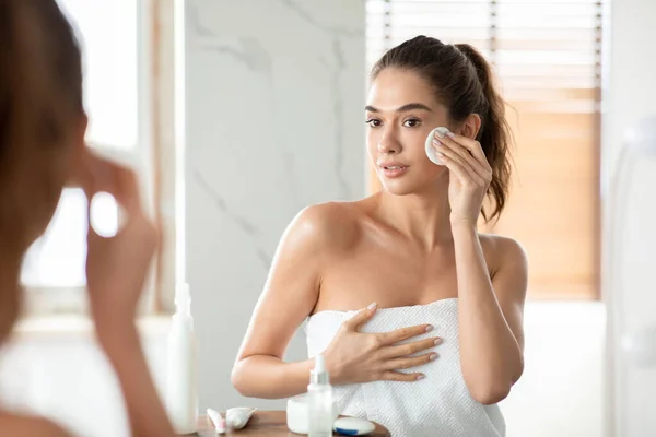 Гарненька жінка використовує бавовняну підкладку для очищення шкіри обличчя у ванній — стокове фото