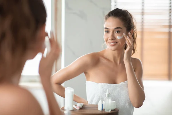 Millennial Lady Applying Facial Moisturizer On Cheek Posing In Bathroom — Stok fotoğraf