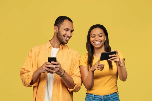 Joyeux jeune couple noir à l'aide de téléphones cellulaires, navigation sur Internet, chat en ligne sur fond de studio jaune — Photo