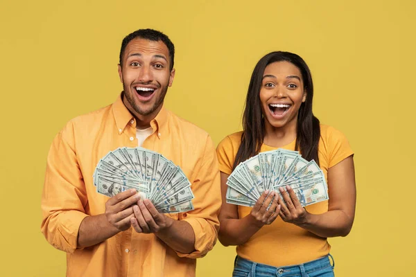 Excited young African American couple holding fans of money, shouting OMG on yellow studio background — Φωτογραφία Αρχείου
