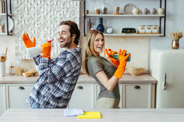 Homem satisfeito com palha em luvas de borracha com spray e esposa cantar no microfone imaginário na cozinha — Fotografia de Stock