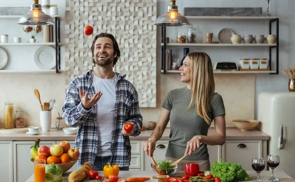 Glad caucasian millennial male with stubble juggles tomatoes, blonde lady prepares salad — Stock fotografie