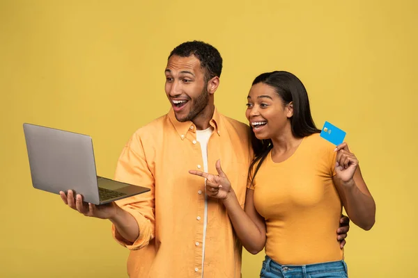 Vzrušený černošky pár nakupování on-line přes notebook a kreditní karty, výherní loterie nebo kasino sázky na žlutém pozadí — Stock fotografie