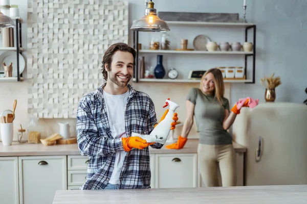 Glad millennial caucasian male with stubble in rubber gloves show spray, wife dust off furniture in kitchen — Stok fotoğraf