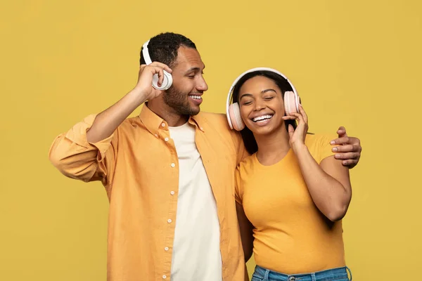 Happy young black couple in wireless headphones listening to music and hugging over yellow studio background — Stock Fotó