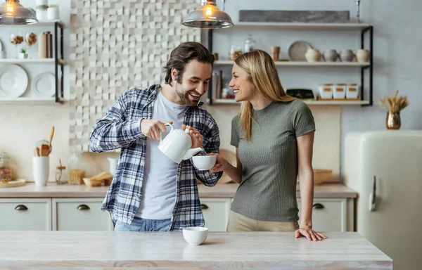 Χαμογελαστός Ευρωπαίος άνδρας χιλιετίας με γένια χύνει τσάι στο φλιτζάνι της ξανθιάς συζύγου στο φως εσωτερικό της κουζίνας — Φωτογραφία Αρχείου