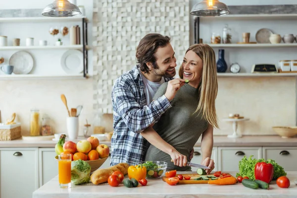 Happy caucasian millennial husband with stubble feeds blonde wife, couple have fun, lady prepare salad — Stock fotografie