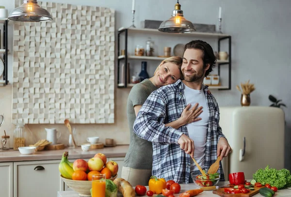 Zâmbind caucazian millennial soție îmbrățișarea om cu mirișcă, tip pregătirea salată la masă — Fotografie, imagine de stoc