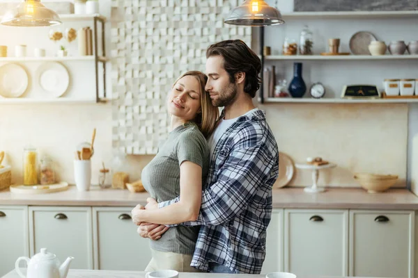 Cheerful happy caucasian millennial hasband with stubble hugs blond wife, enjoy tender moment in light kitchen — Stock fotografie