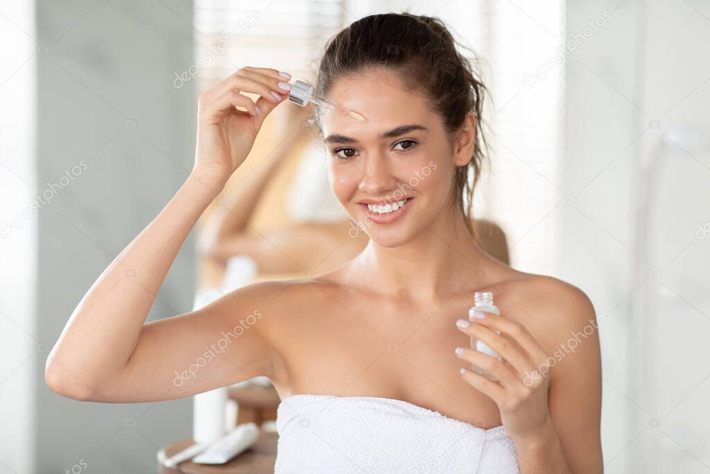 Female Applying Facial Serum On Forehead With Dropper In Bathroom
