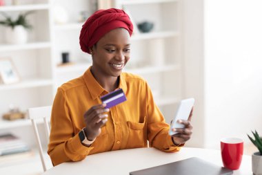 Mutlu Müslüman Afrikalı Amerikalı kadın akıllı telefon ve kredi kartı kullanıyor.