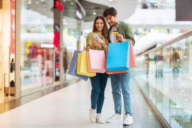 Couple Shopping Using Phone And Credit Card In Mall