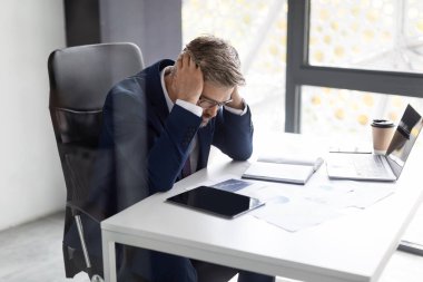 Stressed Mature Businessman In Suit Touching Head While Sitting At Workplace