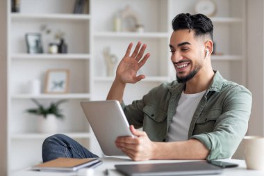 Telsiz kulaklıklı mutlu genç Müslüman adam tablete bakar ve elini sallar.