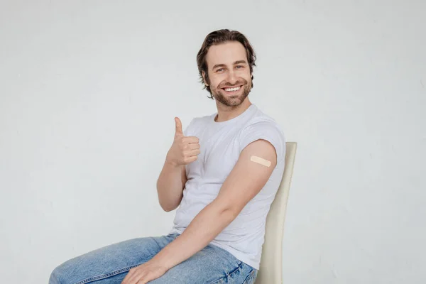 Glad millennial caucasian man in white t-shirt sit on chair, show thumb up and band aid on shoulder — Stockfoto