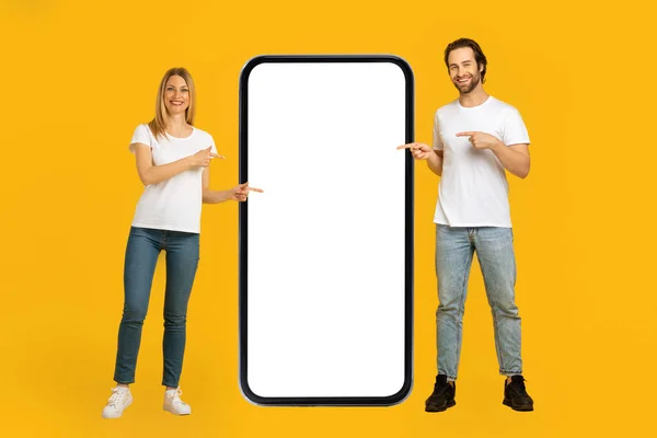Jovem casal europeu alegre em camisetas brancas apontam os dedos para um telefone enorme com tela em branco — Fotografia de Stock