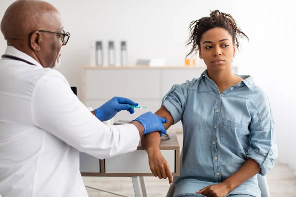 Mujer negra recibiendo la inyección de la vacuna sentada con el médico en el interior —  Fotos de Stock