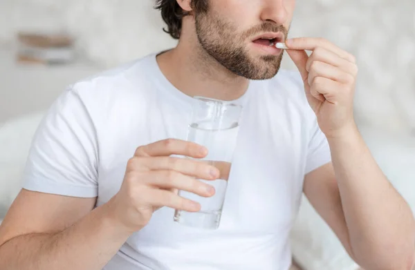 Sad young caucasian man with stubble wake up on white bed, holds glass of water and take pill in bedroom — Stockfoto