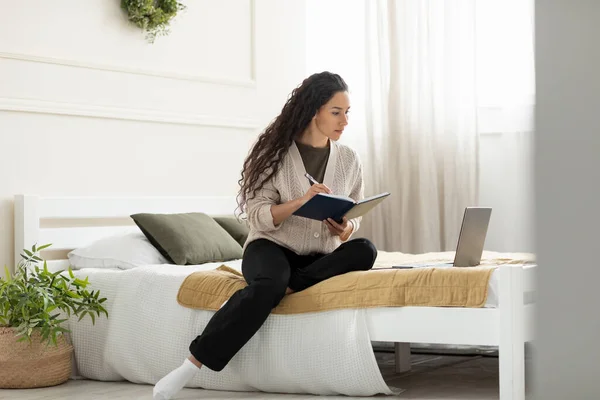 Kvinna som sitter på sängen, använder dator och skriver i anteckningsbok — Stockfoto