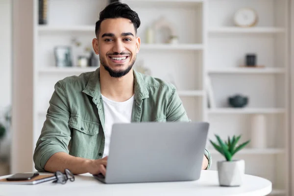 Glad young handsome muslim male with beard looking at camera, working on laptop in home office interior — Stockfoto