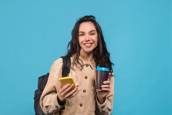 Smiling young european brunette woman with backpack, cup of coffee and smartphone — Stockfoto