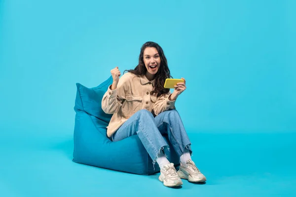 Glad young european brunette female with phone sits on bagchair and makes victory gesture — Stockfoto
