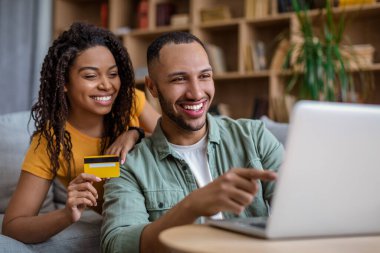 Vacation concept. Happy african american spouses using laptop and credit card, booking tour online, sitting at home