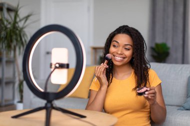 Beauty blogging concept. Happy black lady recording makeup review, using ring lamp and cellphone