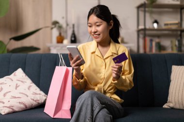 Gülümseyen Asyalı kadın evde telefon ve kredi kartı kullanıyor.