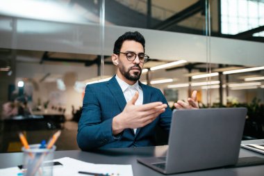 Gözlüklü, takım elbiseli, kablosuz kulaklıklı, laptoplu, ciddi çekici, genç bir Arap.