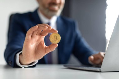 Online Trading. Smiling Businessman In Suit Sitting At Desk And Holding Bitcoin