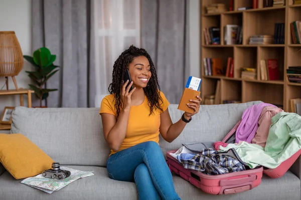 Feliz senhora afro-americana se preparando para férias, falando ao telefone e segurando passaporte com bilhetes, sentado no sofá — Fotografia de Stock