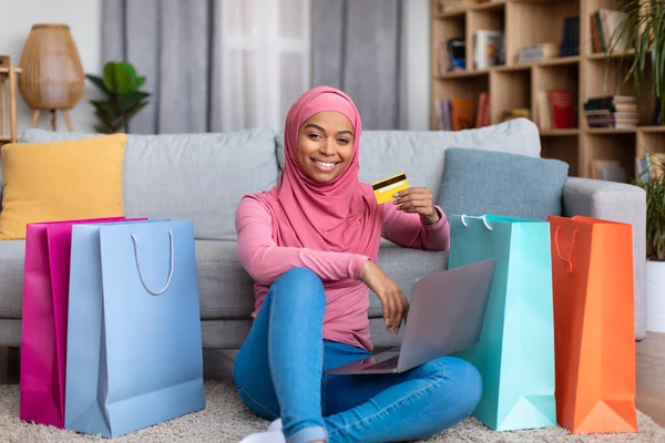 African american woman feeling happy over big sale, using laptop, showing credit card, sitting on floor with gift bags — Stok fotoğraf