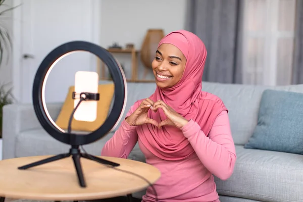 Happy african american female blogger recording video blog on social network, showing the heart symbol with her hands