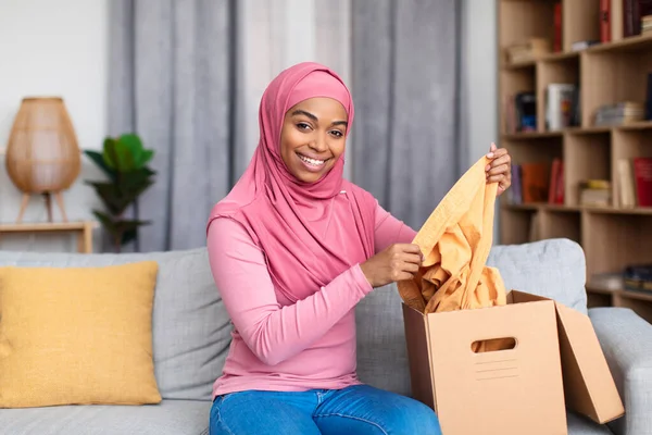 Consegna e shopping online. Felice signora afro-americana in possesso di un pezzo di vestiti, disfare la scatola di cartone — Foto Stock