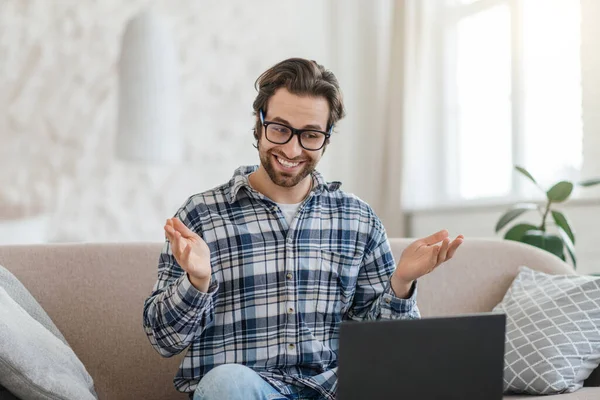 Wesoły młody biały mężczyzna nauczyciel z zarostem w okularach siedzi na kanapie z laptopem i gestykuluje — Zdjęcie stockowe