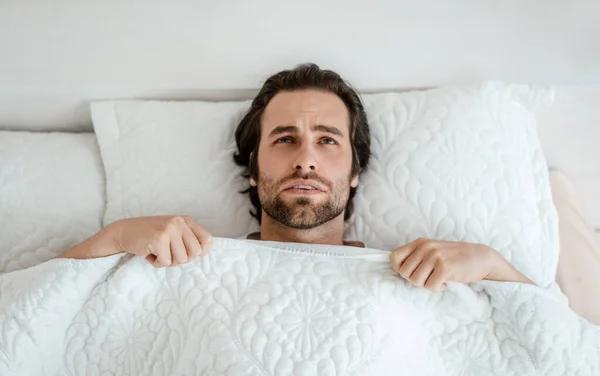 Ongelukkig duizendjarige blanke man met stoppels wakker, slecht gevoel, liggend op wit bed in slaapkamer interieur — Stockfoto