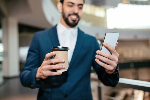 Happy millennial muslim businessman with beard in suit with cup of coffee looks at smartphone in office interior — Zdjęcie stockowe