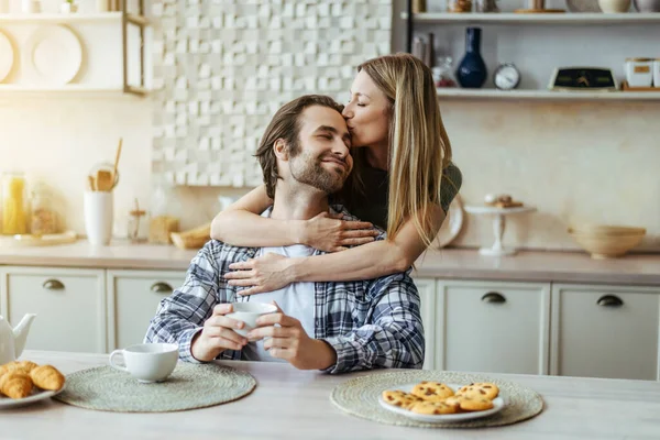 Happy caucasian millennial blonde wife kissing man with stubble on light kitchen interior, sun flare — Stok fotoğraf