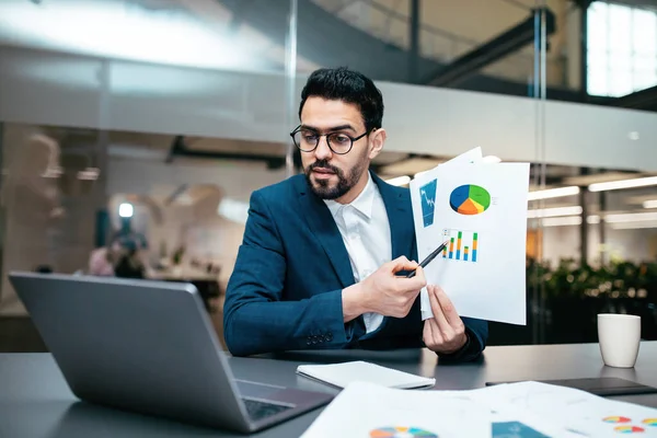 Serious busy millennial arab guy with beard in glasses, suit sits at table shows color chart in computer webcam