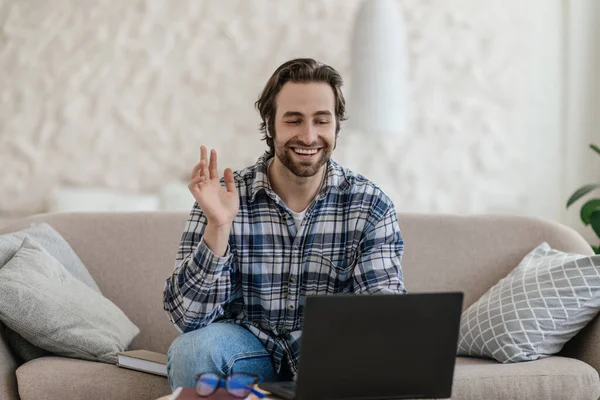 Glad millennial caucasian guy with stubble sits on sofa waving hand and looking at laptop in living room — Foto de Stock