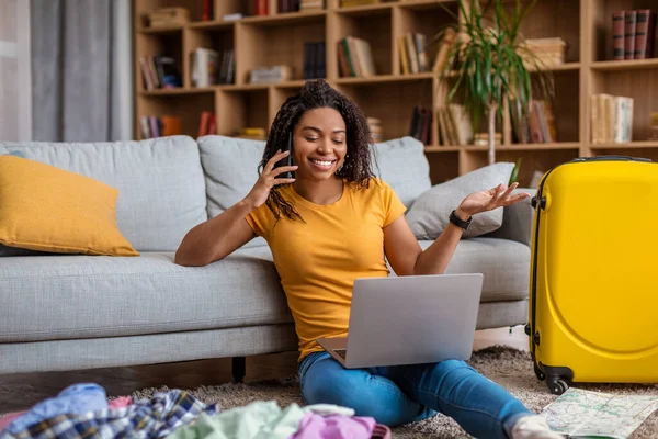 Excitada senhora afro-americana usando laptop e falando ao telefone enquanto procura viagem de verão, se preparando para férias — Fotografia de Stock