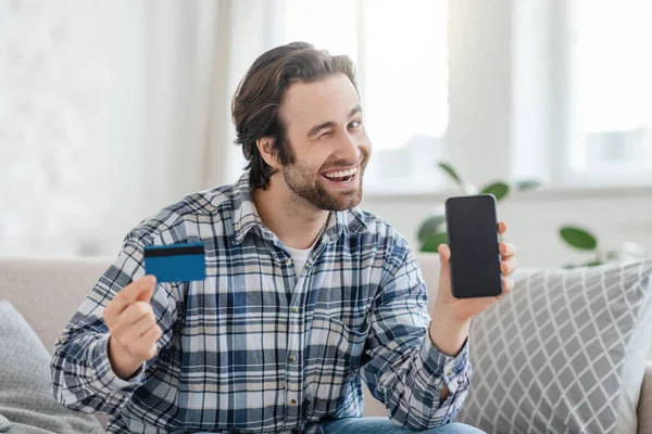 Ικανοποιημένος νεαρός καυκάσιος άνδρας με γένια που κλείνει το μάτι κάθεται στον καναπέ με πιστωτική κάρτα και τηλέφωνο με άδεια οθόνη — Φωτογραφία Αρχείου
