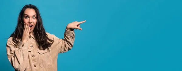 Surprised cheerful young european brunette female closes open mouth with hand, points finger at empty space — Stockfoto